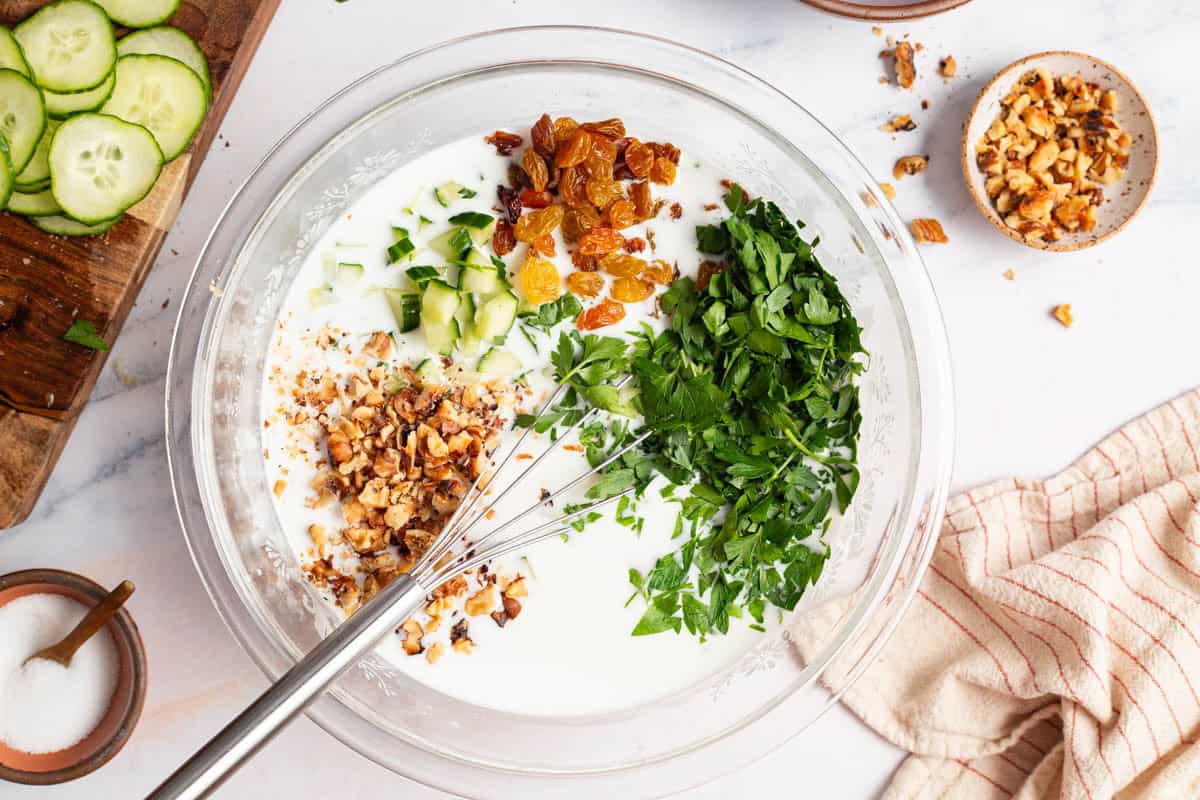 An overhead photo of the parsley, chopped cucumber, golden raisins and walnuts just after being added to the water and yogurt mixture in a bowl with a whisk. Next to this is a kitchen towel, bowls of salt and walnuts, and sliced cucumber on a cutting board.