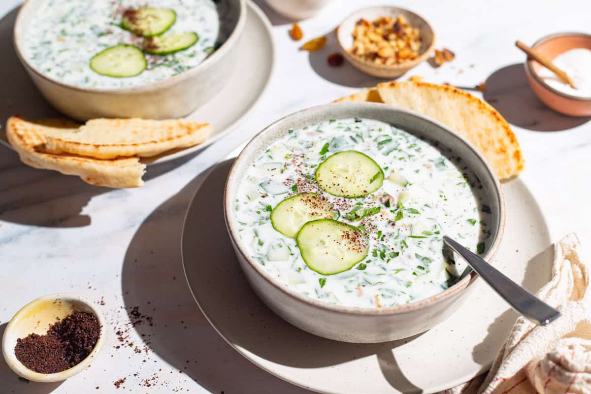 A close up of two bowls of cucumber soup garnished with sumac, parsley and cucumber slices on plates with slices of pita bread, one with a spoon. Next to these are bowls of walnuts, salt and sumac.