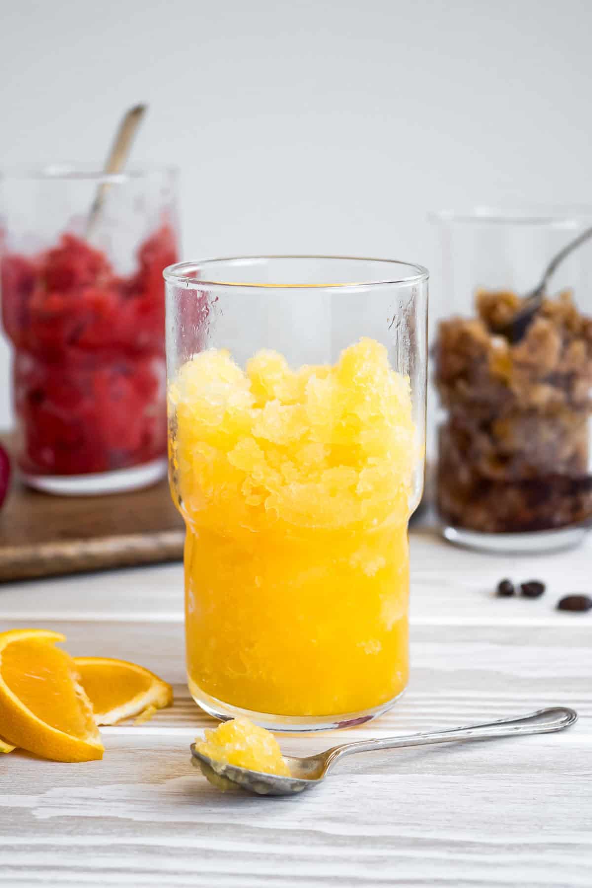 A close up of orange granita in a glass with a spoon and orange slices next to it. In the background is a glass of strawberry granita, and a coffee granita next to some coffee beans.