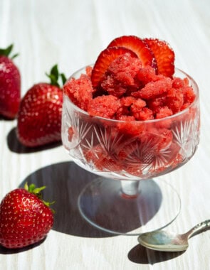 A strawberry granita in a glass garnished with strawberry slices. Next to this is a spoon and 3 strawberries.
