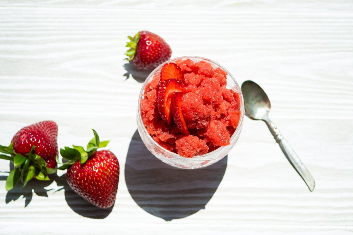 An overhead photo of a strawberry granita in a glass garnished with strawberry slices. Next to this is a spoon and 3 strawberries.