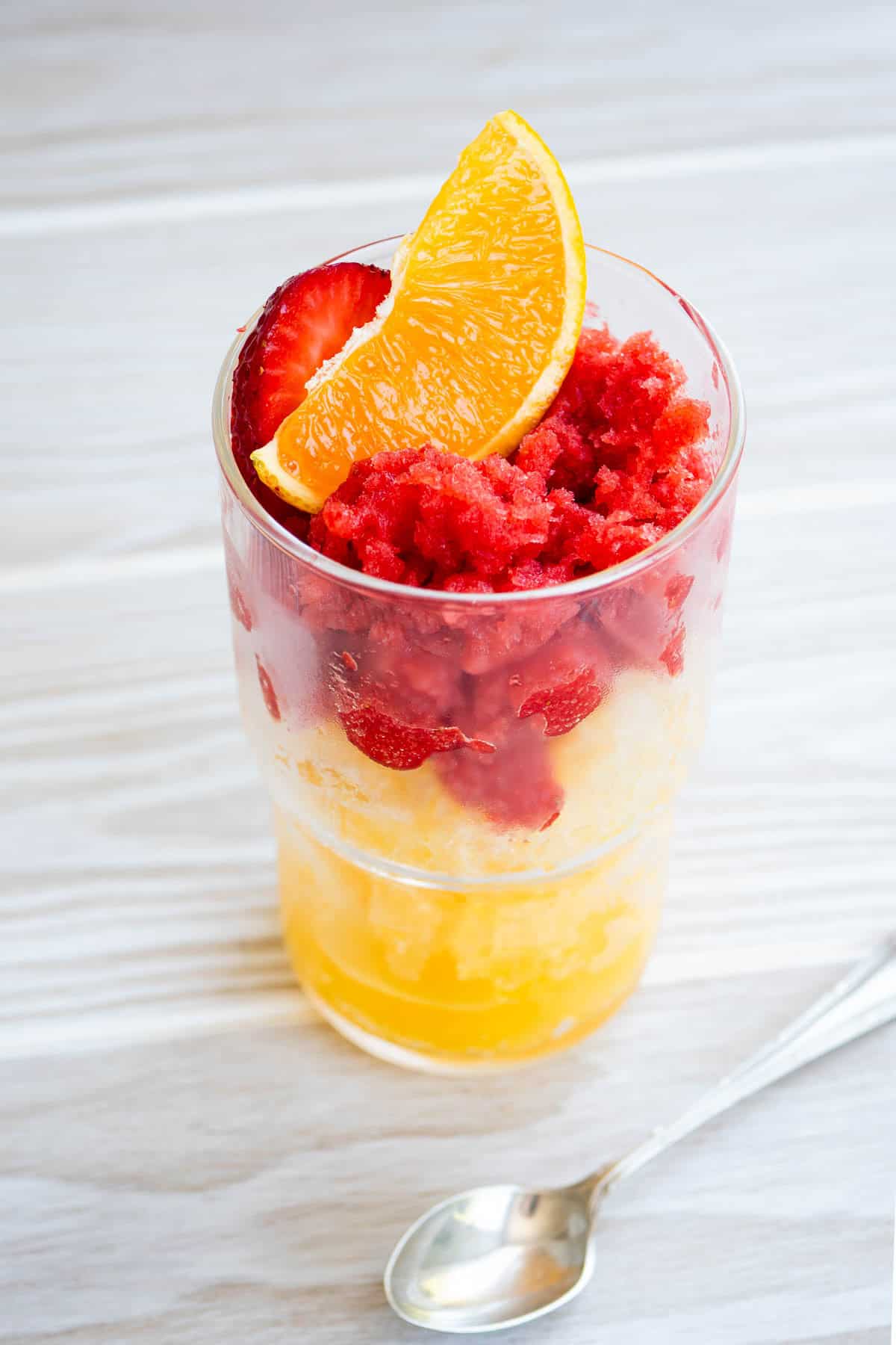 Orange Granita layered with strawberry granita in a glass, garnished with orange and strawberry slices. Next to this is a spoon.