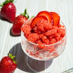 A strawberry granita in a glass garnished with strawberry slices. Next to this is a spoon and 3 strawberries.