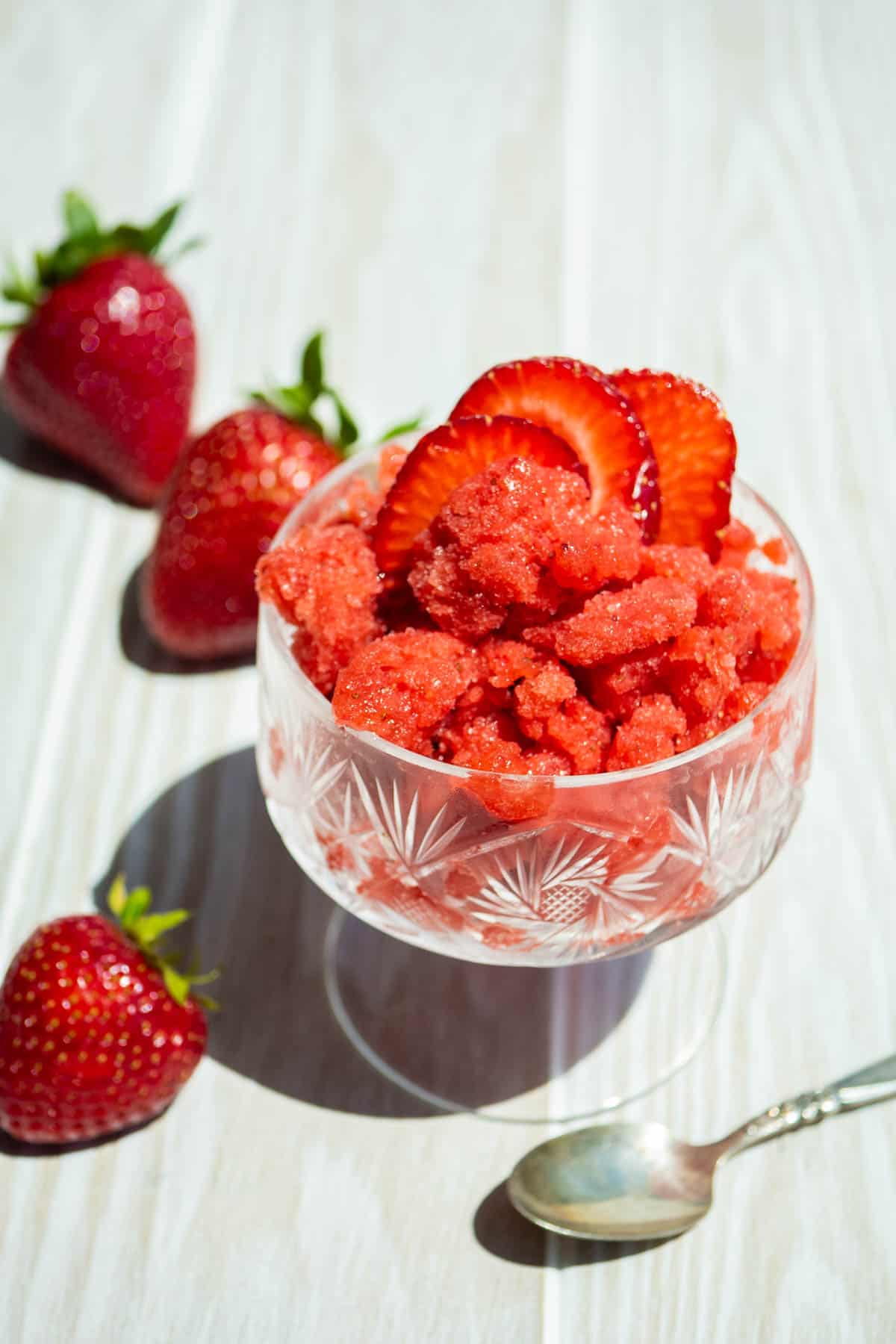 A strawberry granita in a glass garnished with strawberry slices. Next to this is a spoon and 3 strawberries.