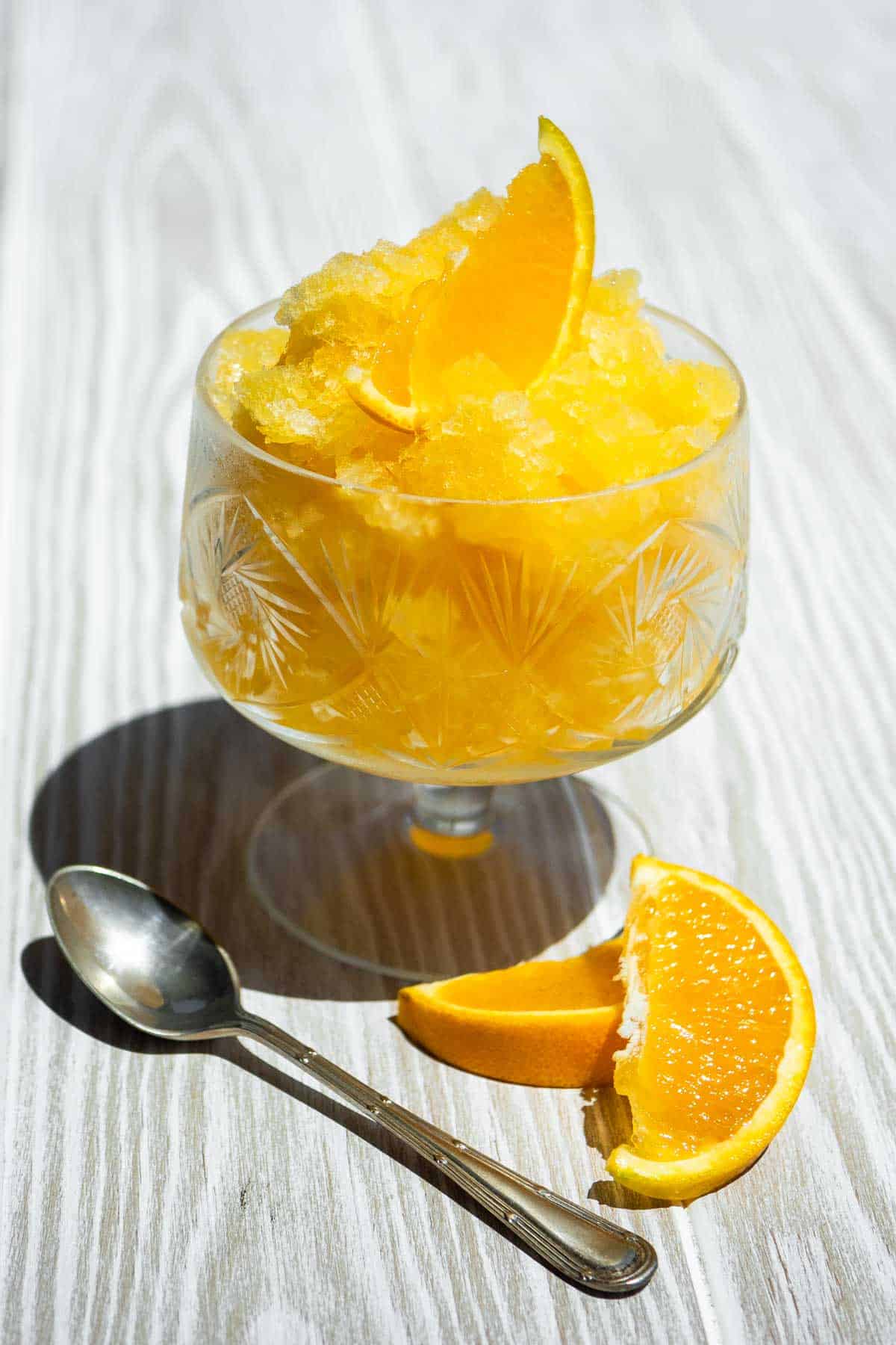 A close up of orange granita garnished with an orange wedge in a bowl next to a spoon and 2 more orange wedges.