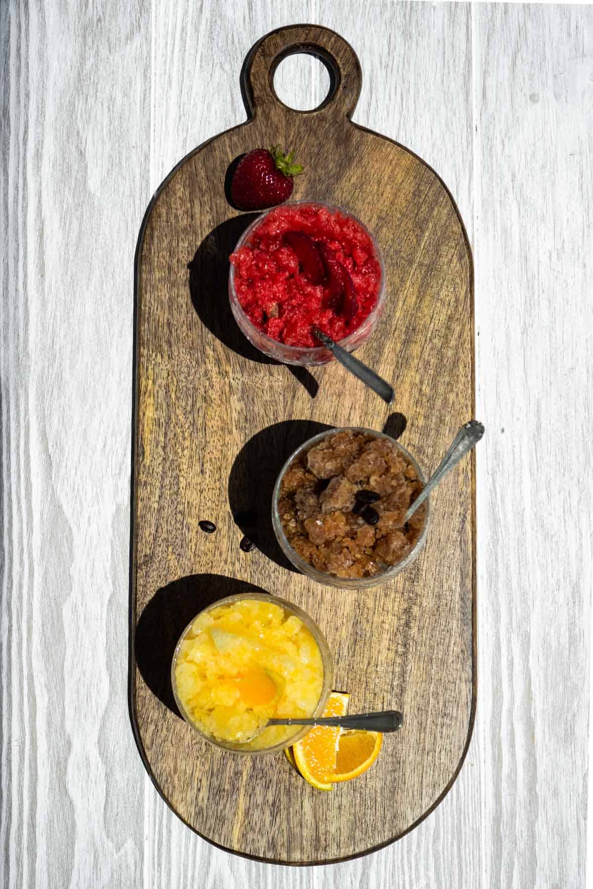 An overhead photo of glasses of strawberry, coffee and orange granita with spoons on a wooden serving tray.