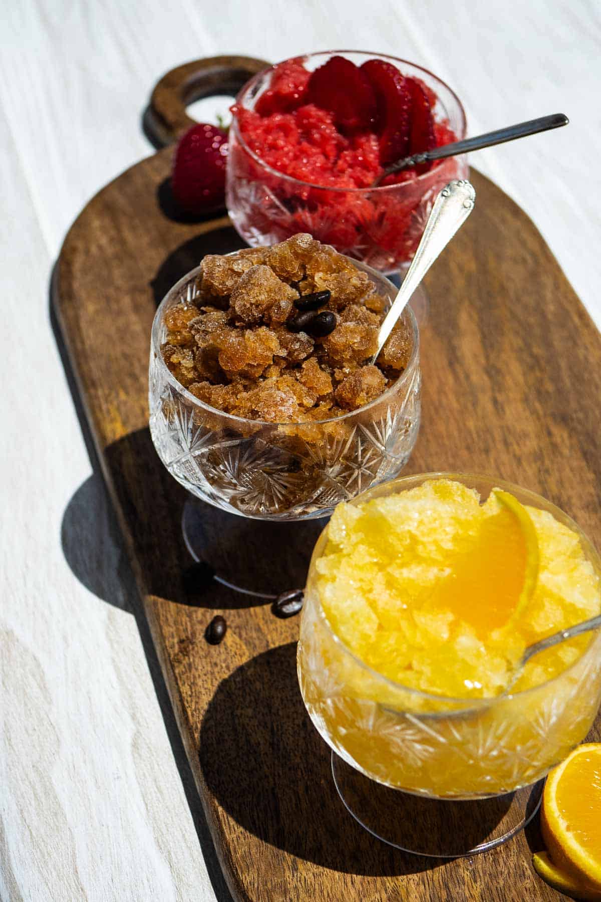 Glasses of strawberry, coffee and orange granita with spoons on a wooden serving tray.