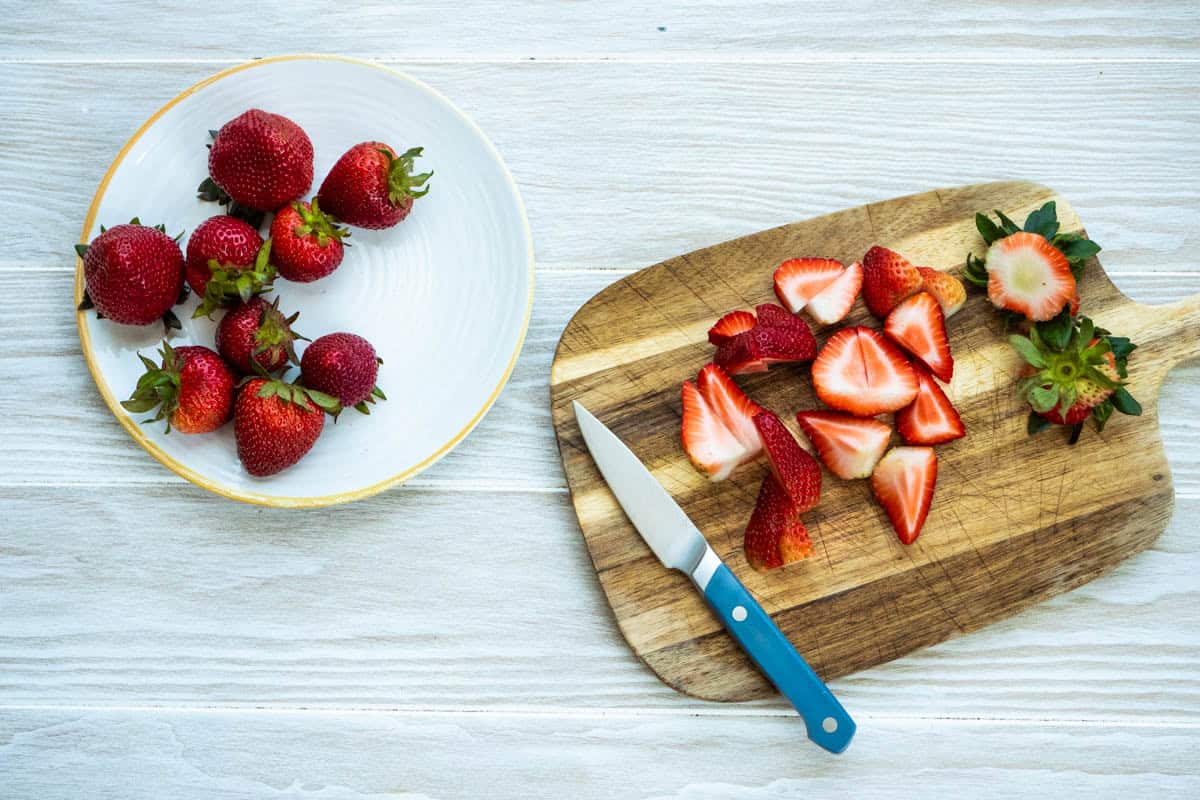 Strawberries on a plate next to a cutting board with pieces of cut up strawberries and a knife.