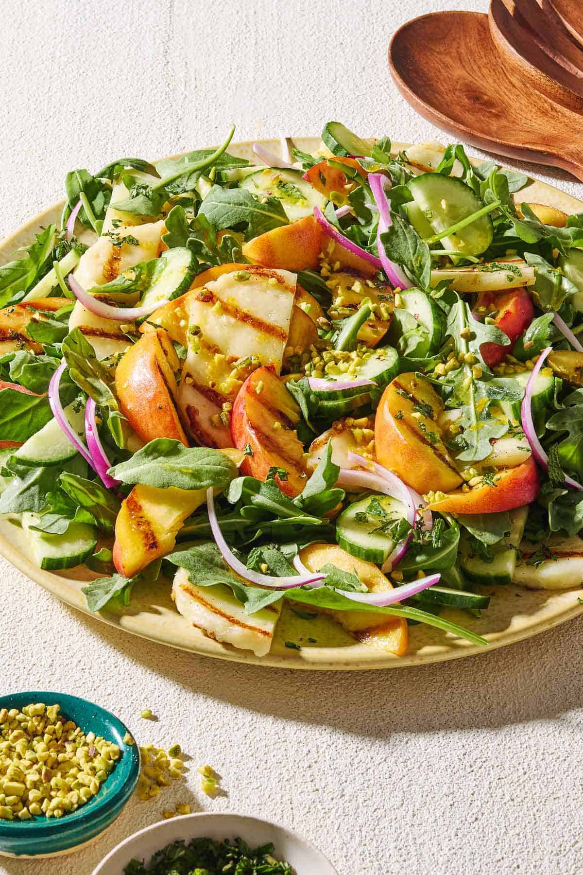 Halloumi and peach salad on a platter next to small bowls of chopped mint leaves and crushed pistachios, and wooden serving utensils.