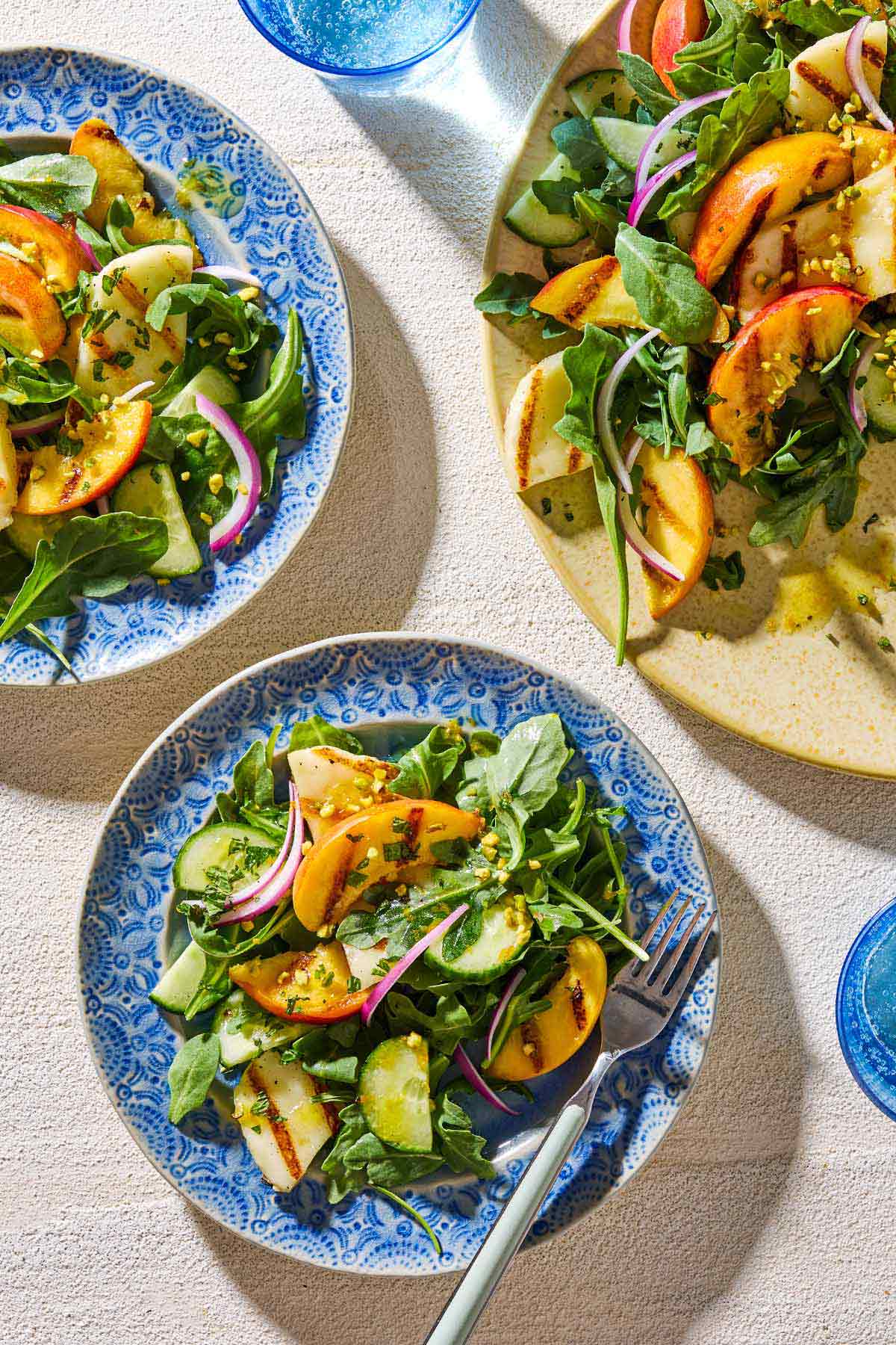 An overhead photo of a serving of halloumi and peach salad on a plate with a fork next to another plate of the salad, the serving platter with the rest of the salad and 2 glasses of water.