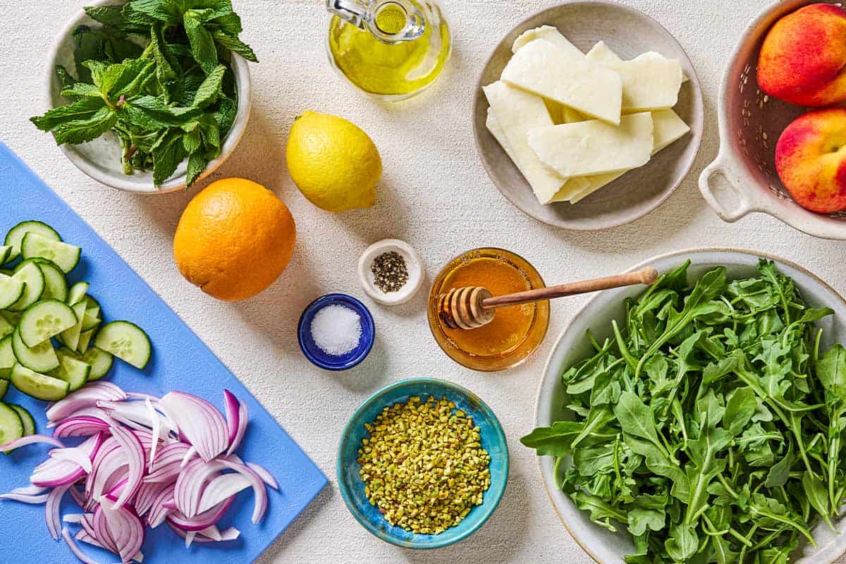 Ingredients for halloumi and peach salad including halloumi, orange, lemon, peaches, olive oil, honey, salt, pepper, mint, red onion, cucumber, pistachios and rocket leaves.