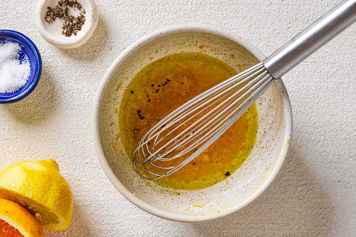 The dressing for the halloumi and peach salad in a bowl with a whisk. Next to this is a lemon half, an orange half and small bowls of salt and pepper.