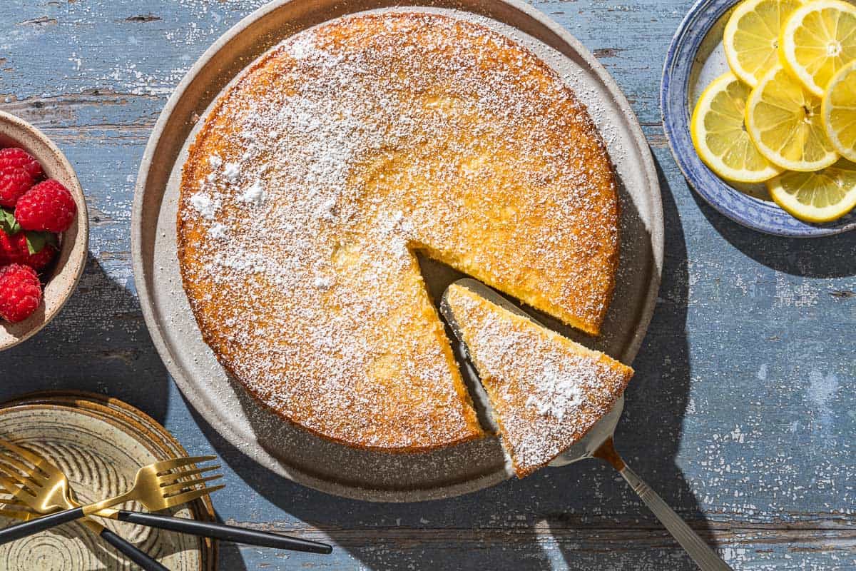 An overhead photo of the lemon ricotta cake on a platter with a slice being removed. Next to this is a plate of lemon slices, a bowl of raspberries, and a stack of plates with 3 forks.