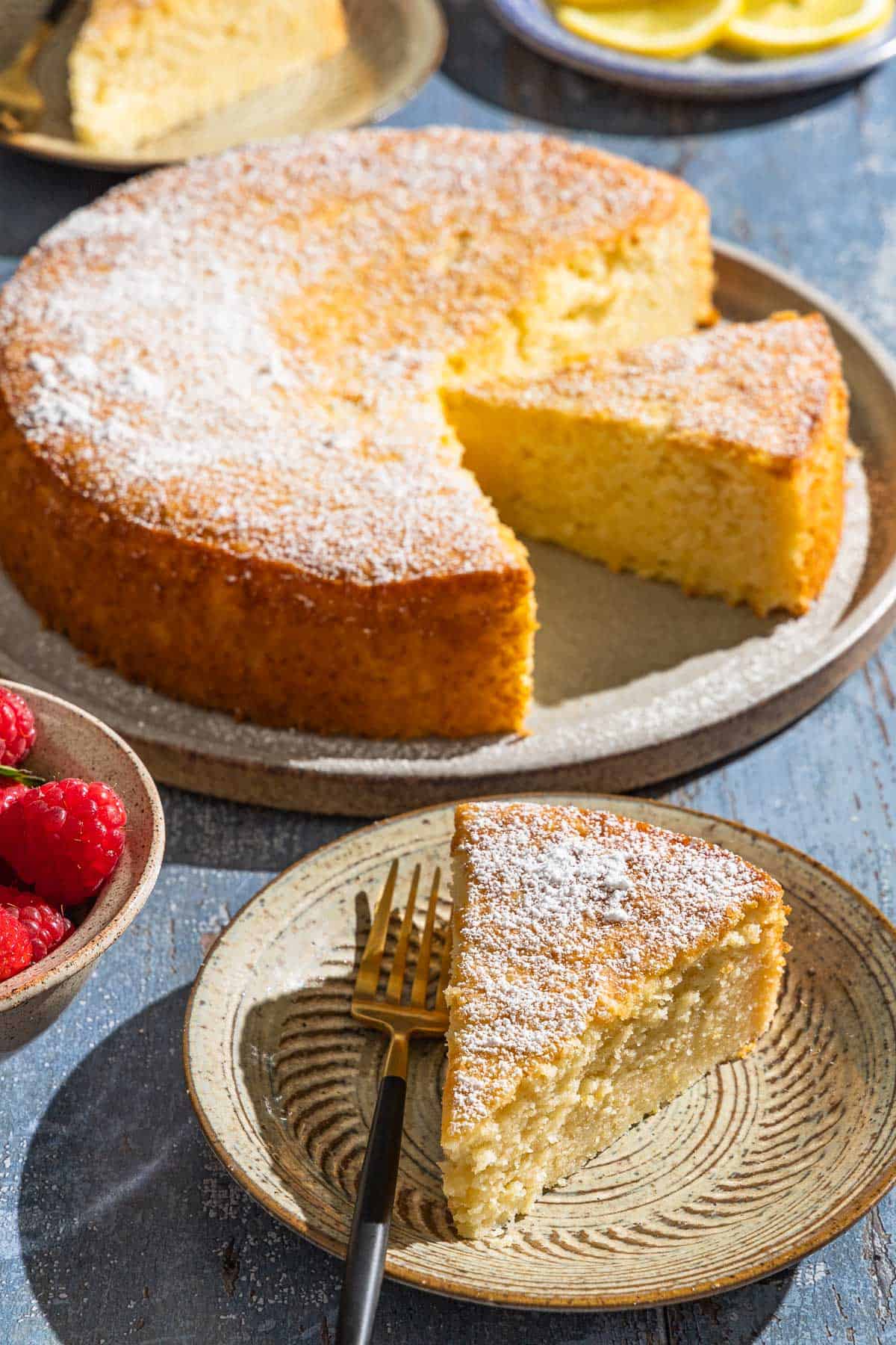 A slice of lemon ricotta cake on a plate with a fork in front of a bowl of raspberries and the rest of the cake on a serving platter.