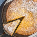 An overhead photo of the lemon ricotta cake on a platter with a slice being removed. Next to this is a plate of lemon slices.