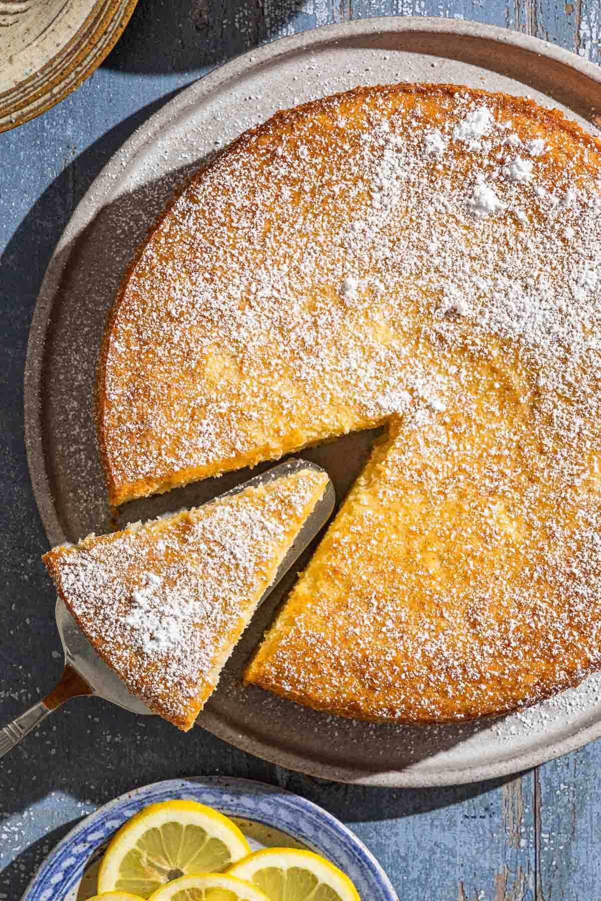 An overhead photo of the lemon ricotta cake on a platter with a slice being removed. Next to this is a plate of lemon slices.