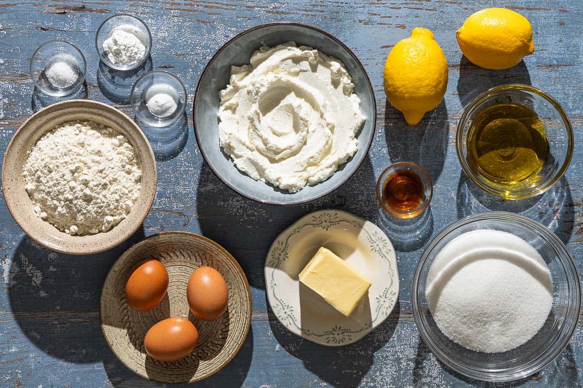 Ingredients for lemon ricotta cake including flour, baking powder, baking soda, salt, butter, olive oil, sugar, lemons, eggs, ricotta cheese, vanilla extract and powdered sugar.