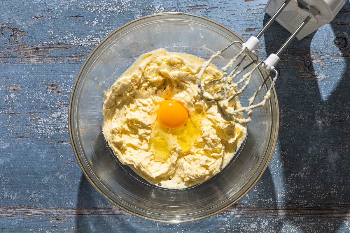 The ingredients for the lemon ricotta cake combined in a mixing bowl just before being mixed together with an egg. Sitting next to this is a hand mixer.