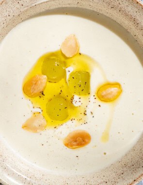 An overhead photo of ajo blanco white gazpacho garnished with green grapes, toasted almonds, olive oil and black pepper with a spoon next to it.