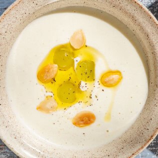 An overhead photo of ajo blanco white gazpacho garnished with green grapes, toasted almonds, olive oil and black pepper with a spoon next to it.