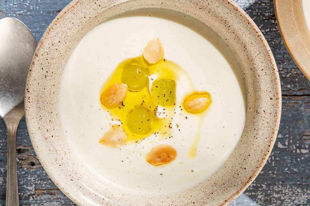 An overhead photo of ajo blanco white gazpacho garnished with green grapes, toasted almonds, olive oil and black pepper with a spoon next to it.