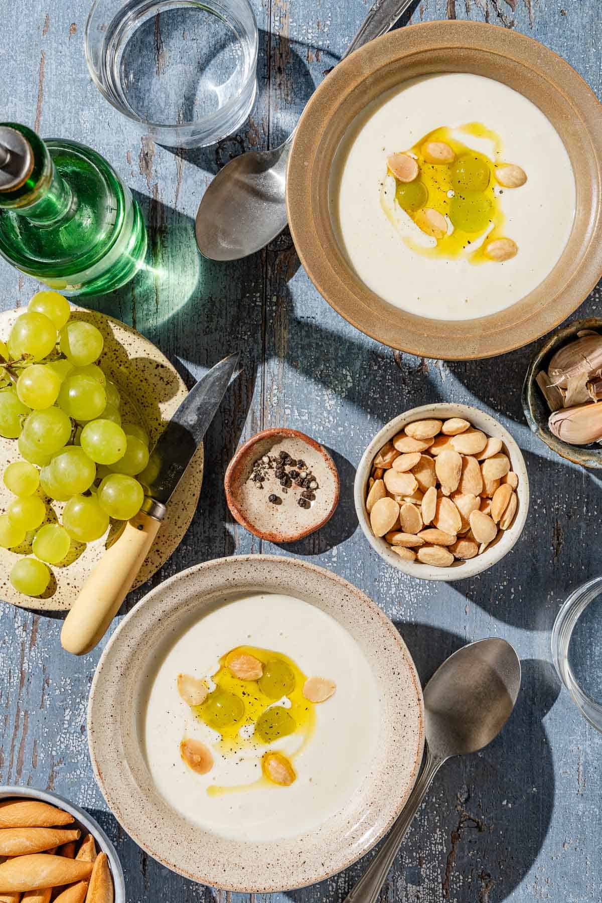 An overhead photo of 2 bowls of ajo blanco white gazpacho garnished with green grapes, toasted almonds, olive oil and black pepper. Next to these are spoons, bowls of almonds, garlic and black pepper, a plate of grapes with a knife, cups of water, and bottle of olive oil.