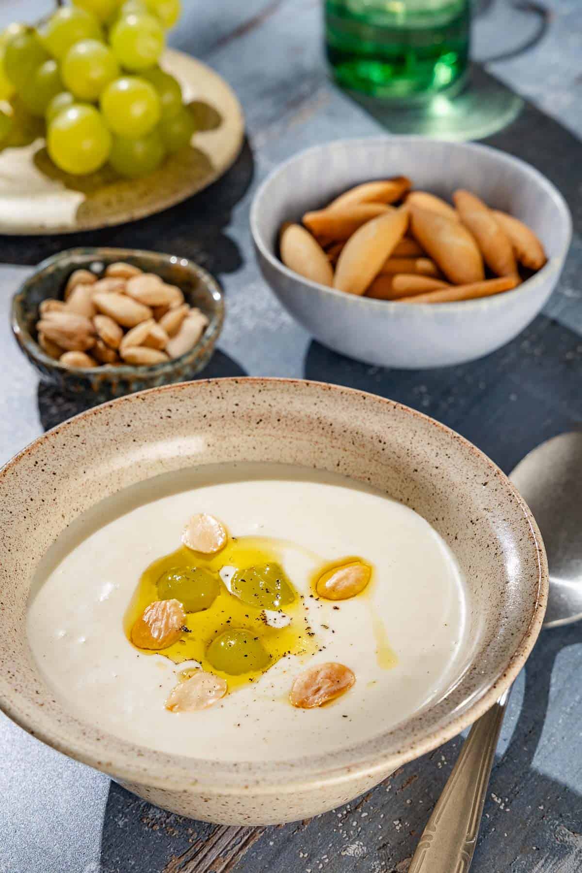 A close up of ajo blanco white gazpacho garnished with green grapes, toasted almonds, olive oil and black pepper. Next to this is a spoon, bowls of almonds and a plate of grapes.