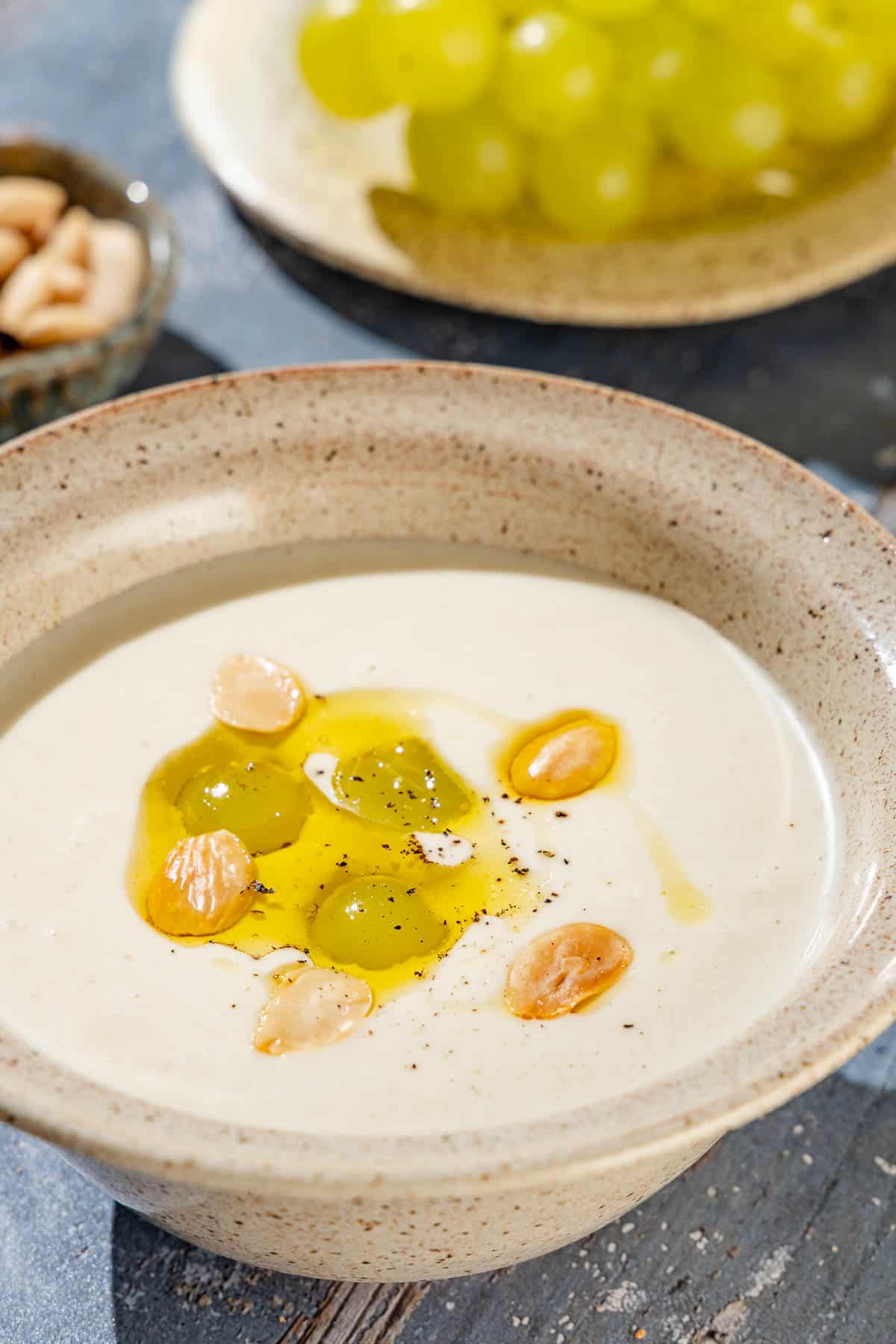 A close up of ajo blanco white gazpacho garnished with green grapes, toasted almonds, olive oil and black pepper next to a plate of grapes.