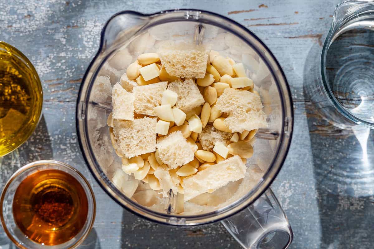 An overhead photo of cubes of bread, blanched almonds and garlic in the pitcher of a blender next to cups of olive oil, sherry vinegar, and water.