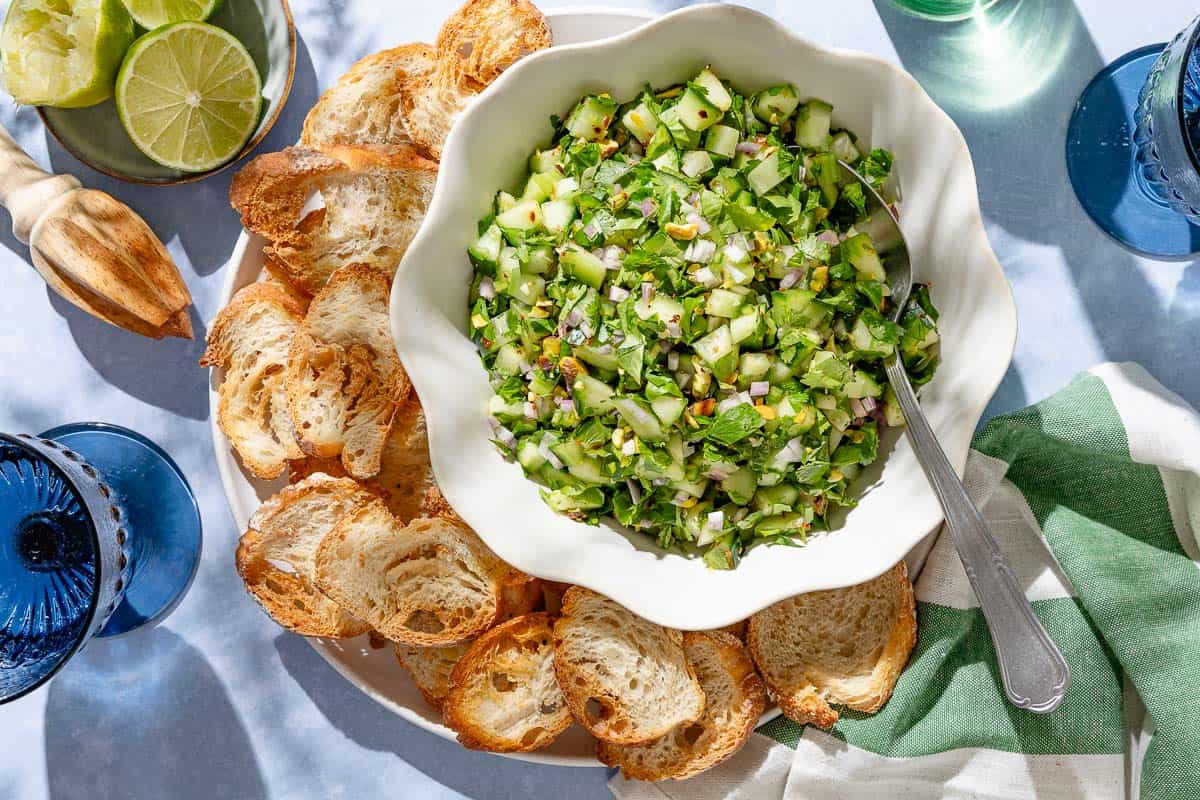 An overhead photo of cucumber salsa in a bowl with a spoon on a platter with slices of toasted baguette. Next to this is a kitchen towel, a plate of limes , a juicer and two glasses of water.