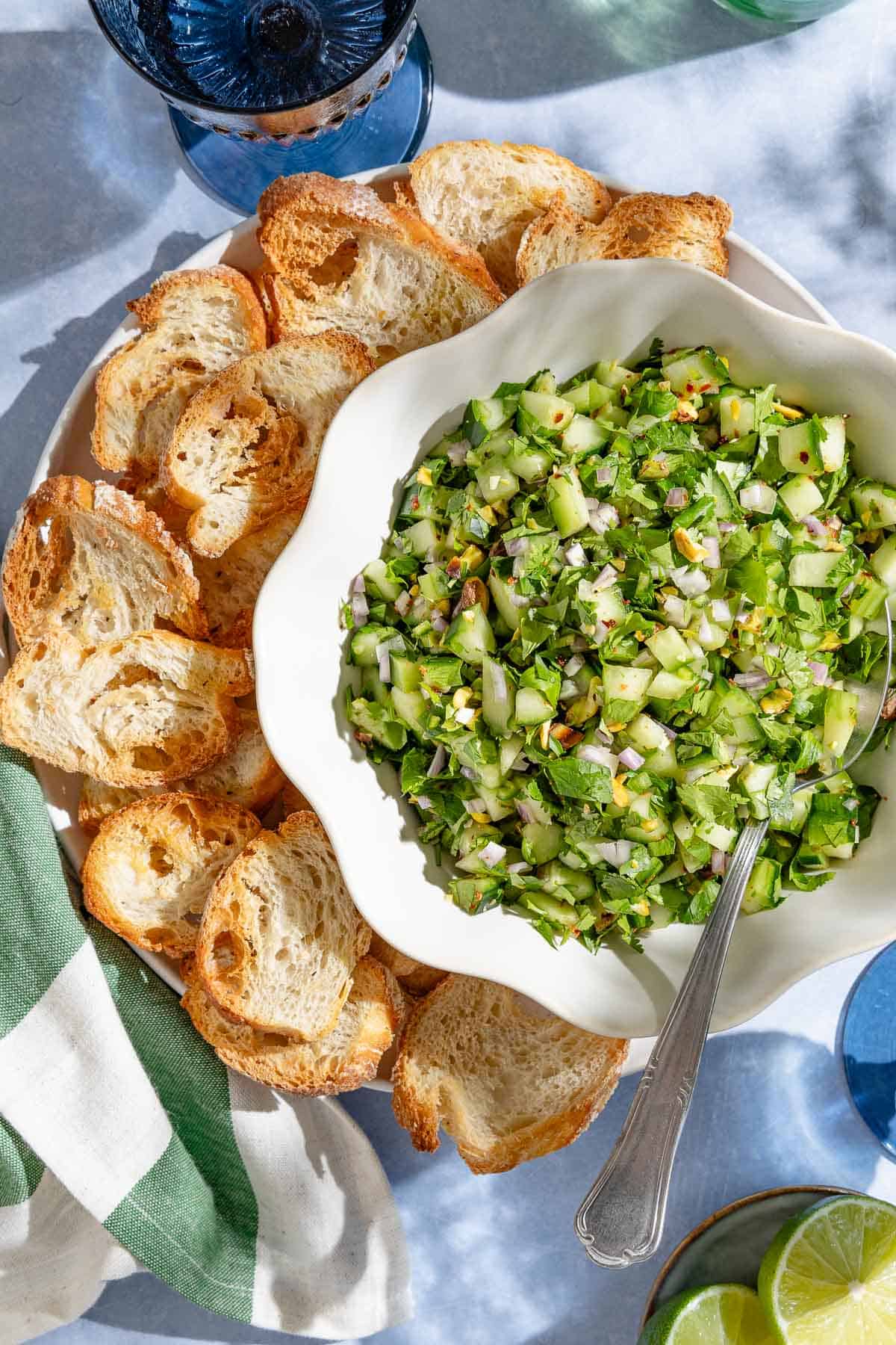 An overhead photo of cucumber salsa in a bowl with a spoon on a platter with slices of toasted baguette. Next to this is a kitchen towel, a plate of limes and a glass of water.