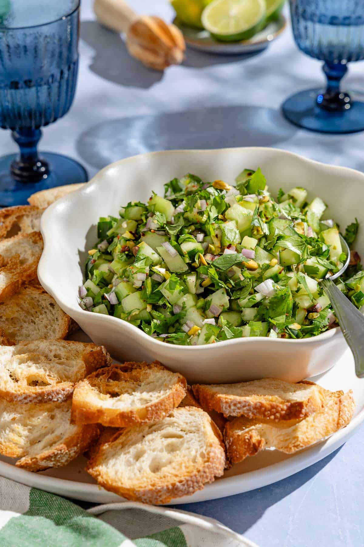 Cucumber salsa in a bowl with a spoon on a platter with slices of toasted baguette. Next to this is a kitchen towel, a plate of limes and two glasses of water.