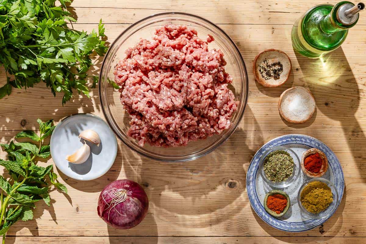 Ingredients for greek style lamb burger recipe including ground lamb, red onion, garlic, fresh parsley, mint, oregano, cumin, paprika cayenne pepper, salt, black pepper, and olive oil.