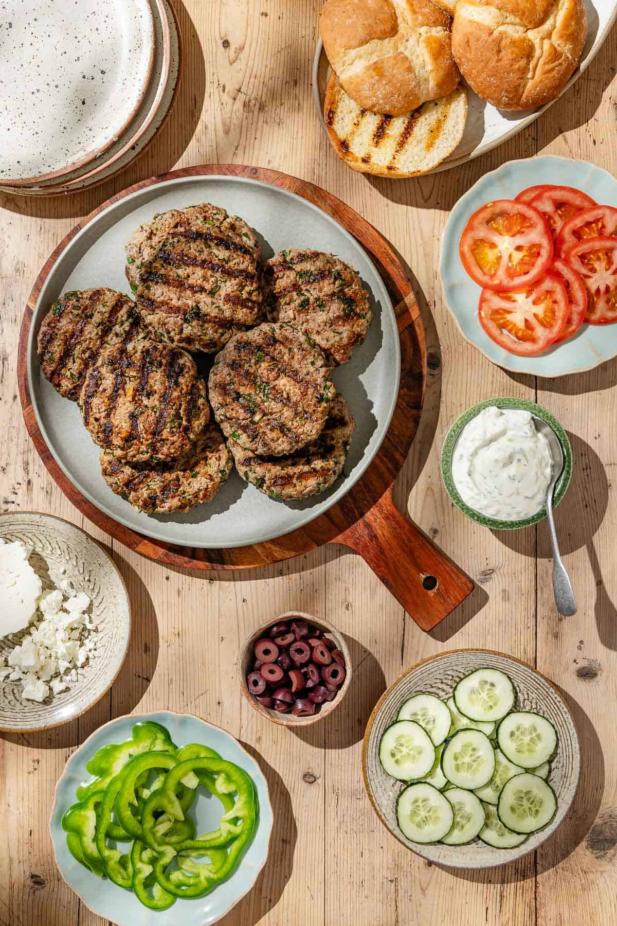 An overhead photo of grilled greek style lamb burger patties on a serving platter on a wooden try. Surrounding this is a stack of plates, plates of feta cheese, sliced cucumbers, sliced tomatoes, sliced green peppers and grilled buns as well as bowls of kalamata olives and tzatziki sauce.