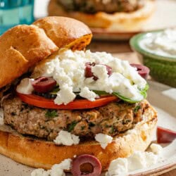 A close up of a greek style lamb burger on a plate in front of various table settings.