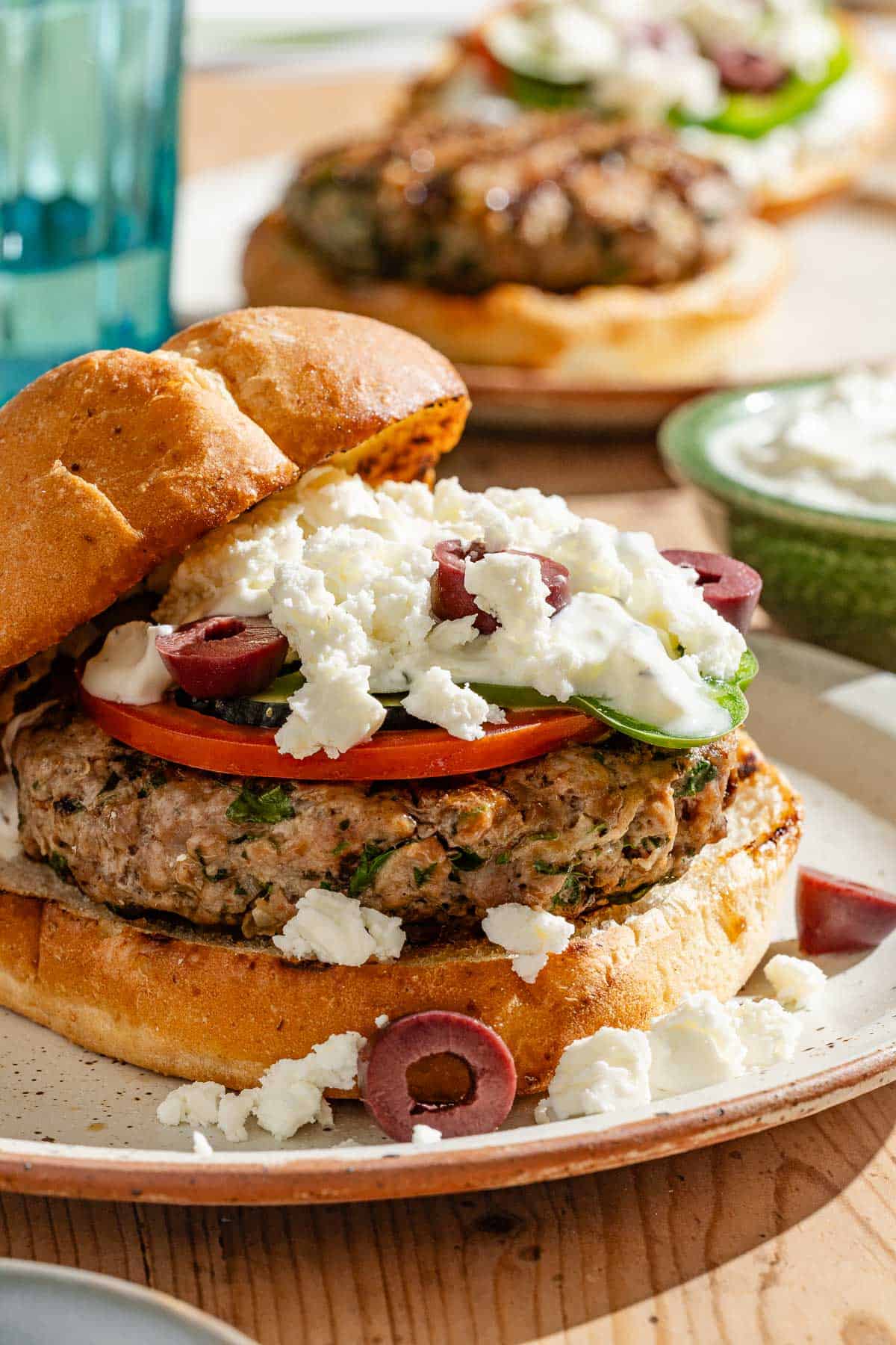 A close up of a greek style lamb burger on a plate in front of various table settings.