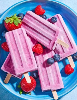 An overhead photo of 4 yogurt berry homemade popsicles surrounded by blueberries, strawberries and raspberries on a plate.
