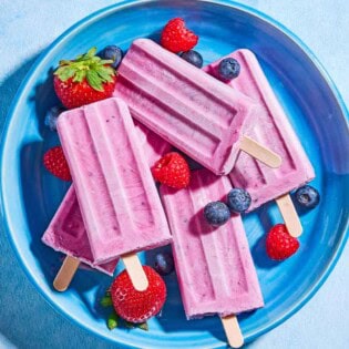 An overhead photo of 4 yogurt berry homemade popsicles surrounded by blueberries, strawberries and raspberries on a plate.