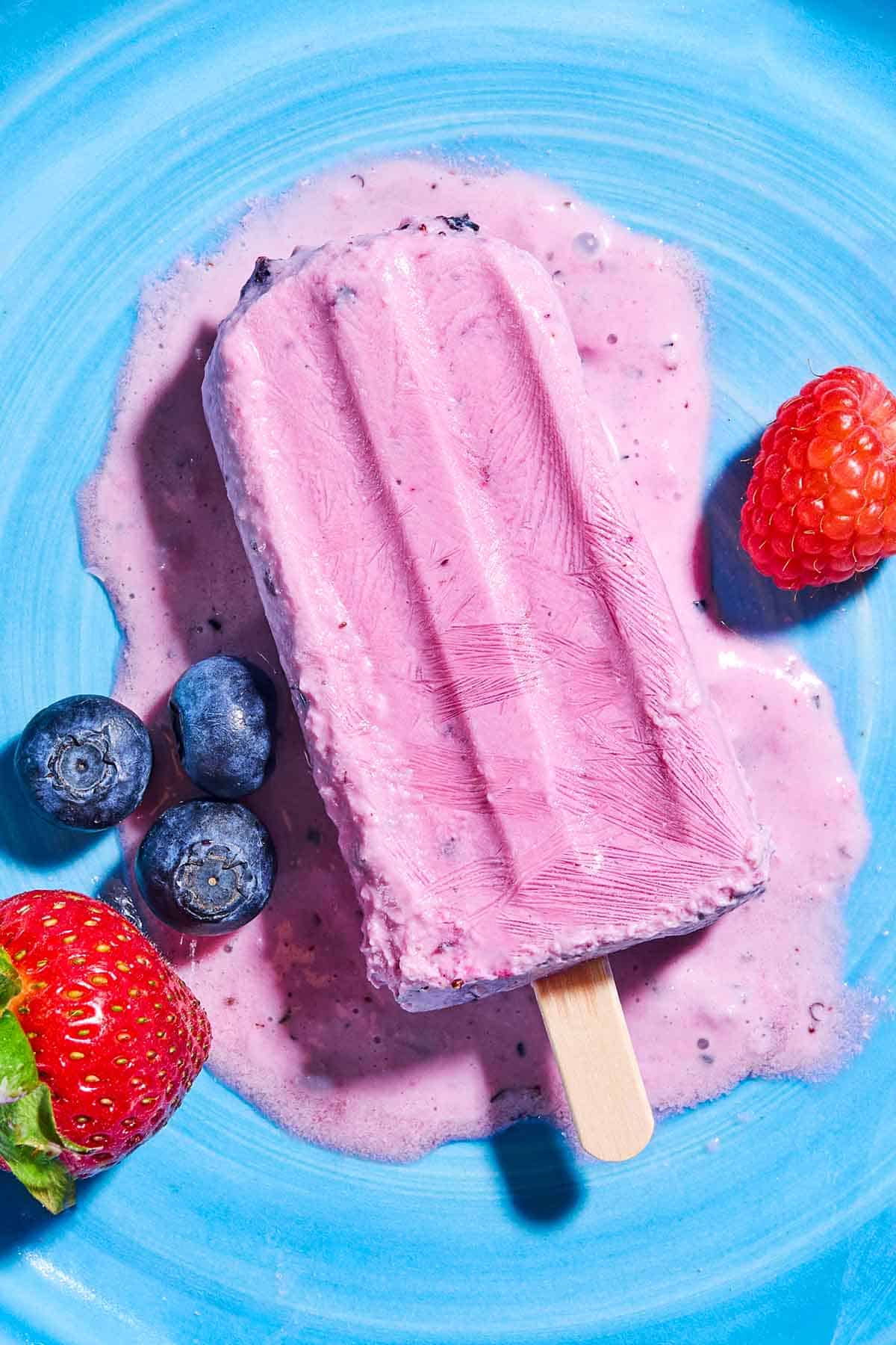 An overhead photo of a slightly melted yogurt berry homemade popsicle on a plate surrounded by blueberries, a strawberry and a raspberry.