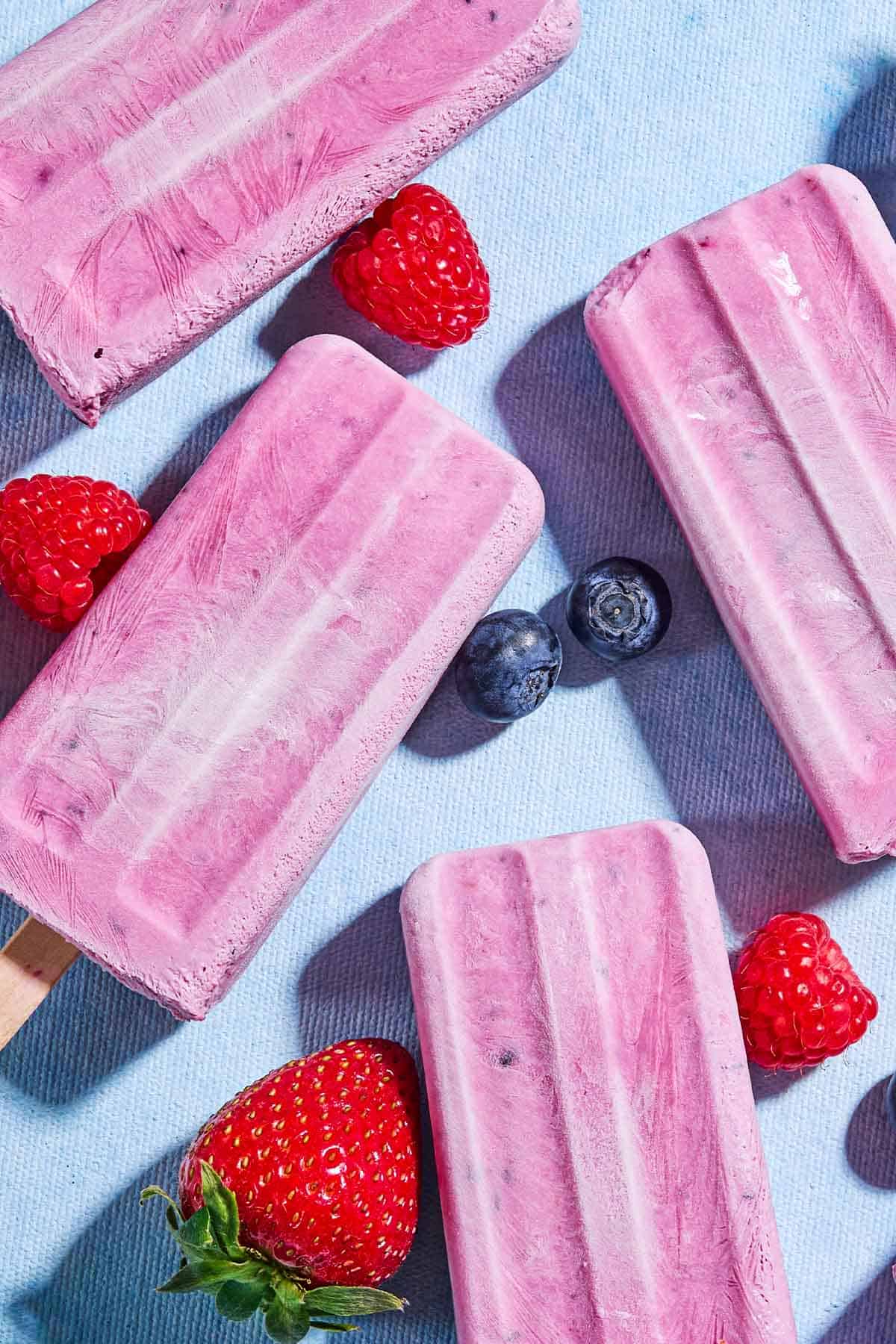 An overhead close up photo of 4 yogurt berry homemade popsicles surrounded by blueberries, strawberries and raspberries.