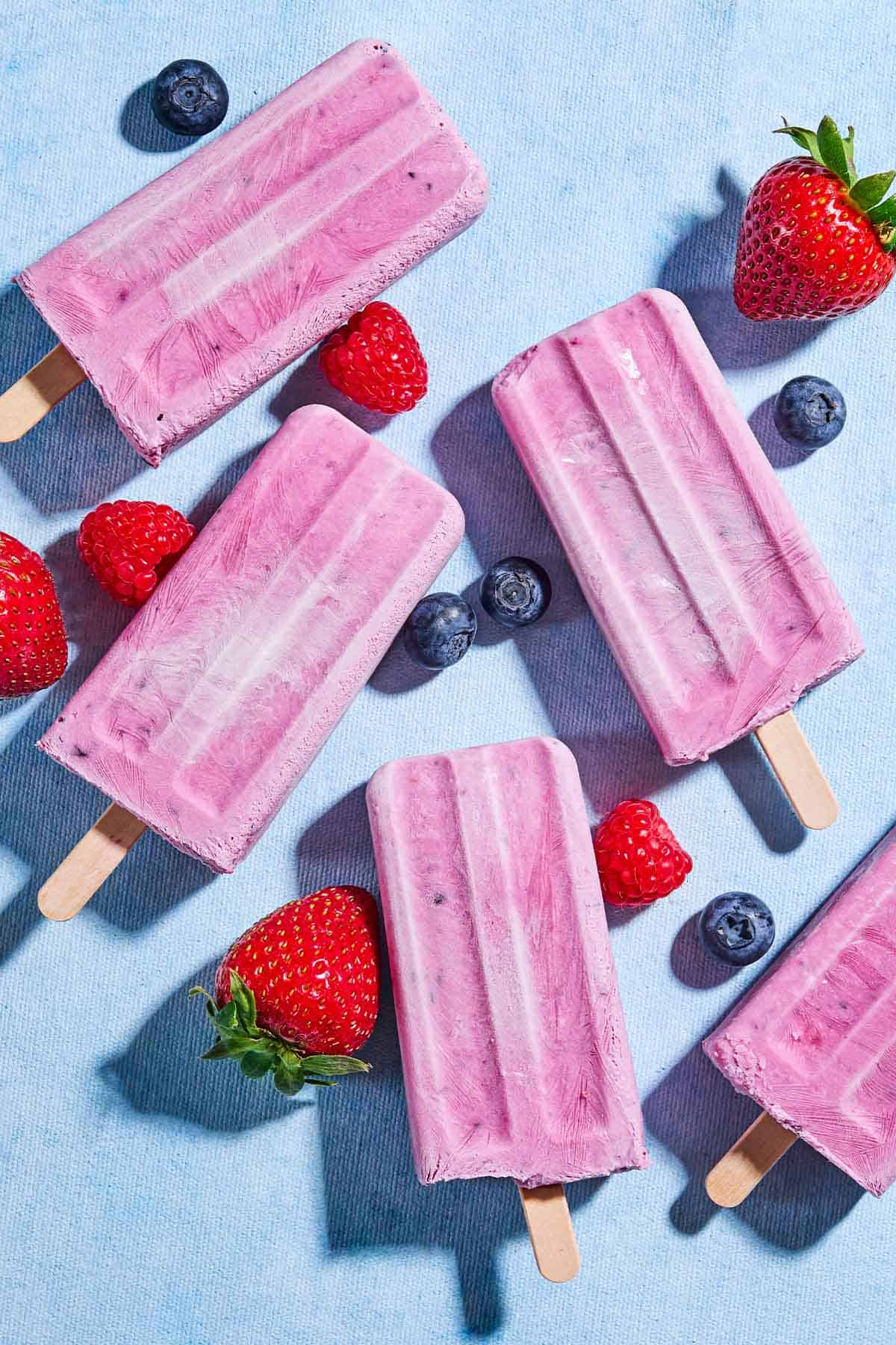 An overhead photo of 4 yogurt berry homemade popsicles surrounded by blueberries, strawberries and raspberries.