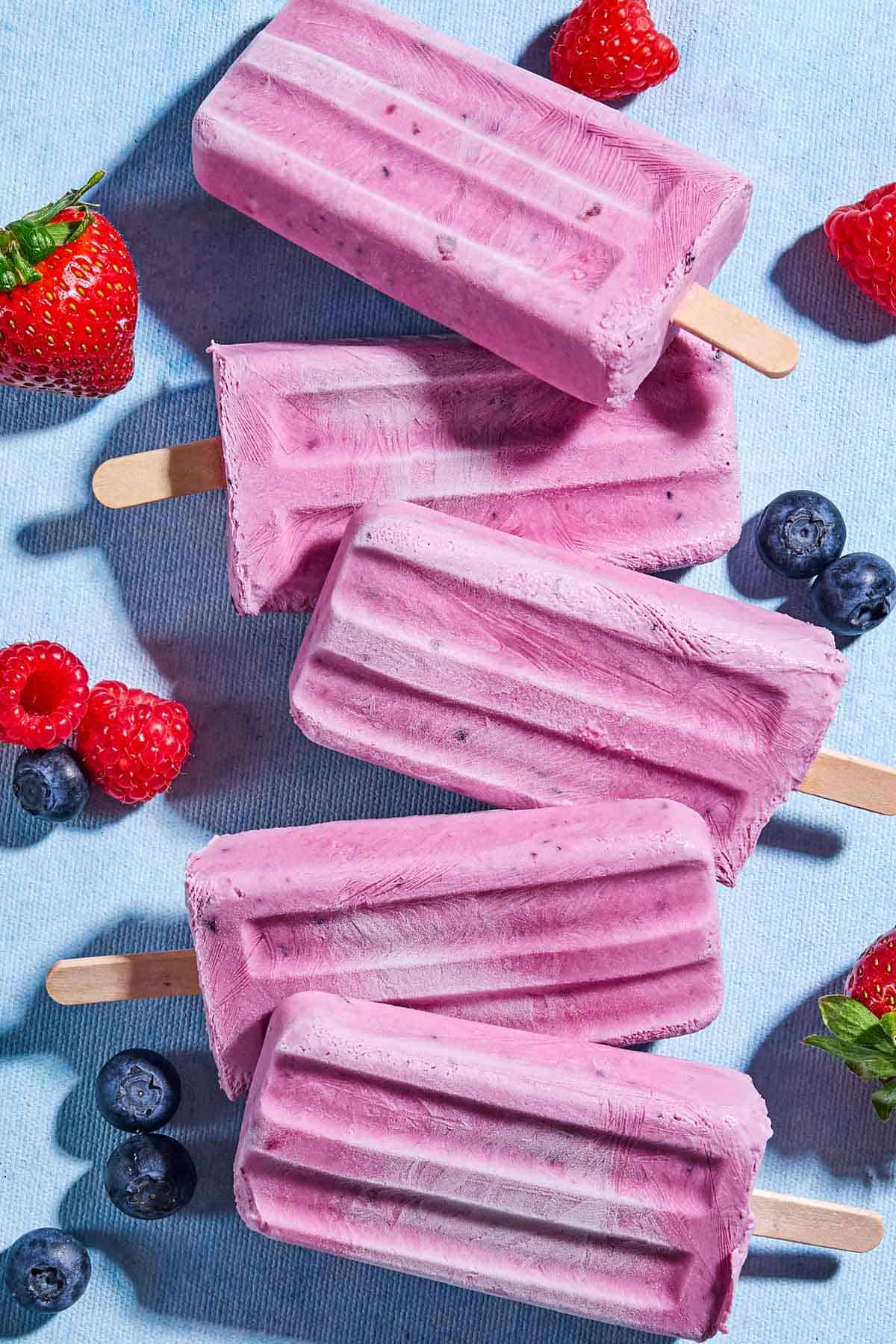 An overhead photo of 5 yogurt berry homemade popsicles surrounded by blueberries, strawberries and raspberries.