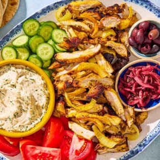 An overhead photo of the sliced grilled chicken shawarma, cucumbers, tomatoes, and bowls of tzatziki, kalamata olives and pickled onions on a serving platter. Next to this is a stack of pita bread.