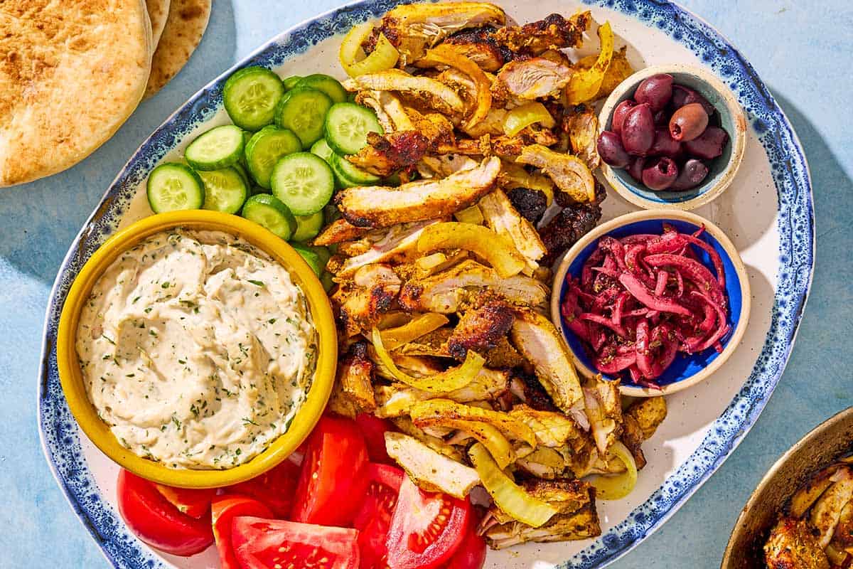 An overhead photo of the sliced grilled chicken shawarma, cucumbers, tomatoes, and bowls of tzatziki, kalamata olives and pickled onions on a serving platter. Next to this is a stack of pita bread.