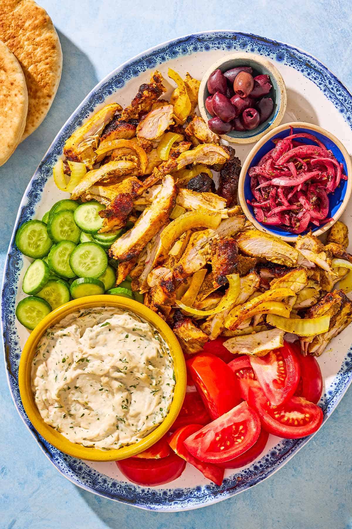 An overhead photo of the sliced grilled chicken shawarma, cucumbers, tomatoes, and bowls of tzatziki, kalamata olives and pickled onions on a serving platter. Next to this is a stack of pita bread.