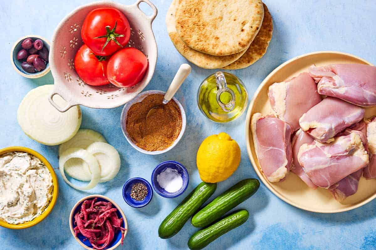 Ingredients for grilled chicken shawarma including chicken thighs, shawarma spice blend, olive oil, lemon, salt, pepper, onion. tomatoes, persian cucumbers, pickled onions, kalamata olives, tahini sauce, and pita bread.