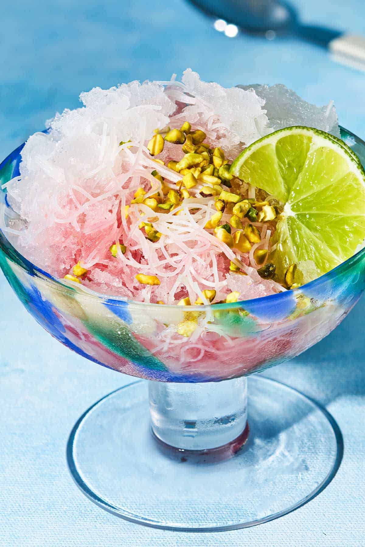 A close up of the faloodeh in a bowl topped with the cherry syrup, chopped pistachios and lime wedges.