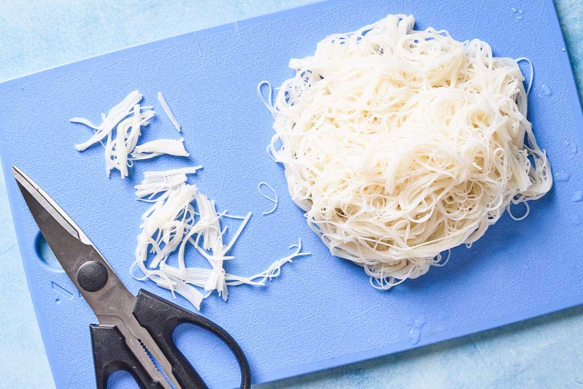 The cooked rice vermicelli on a cutting board with a pair of kitchen shears. Some of the vermicelli has been cut into small pieces.
