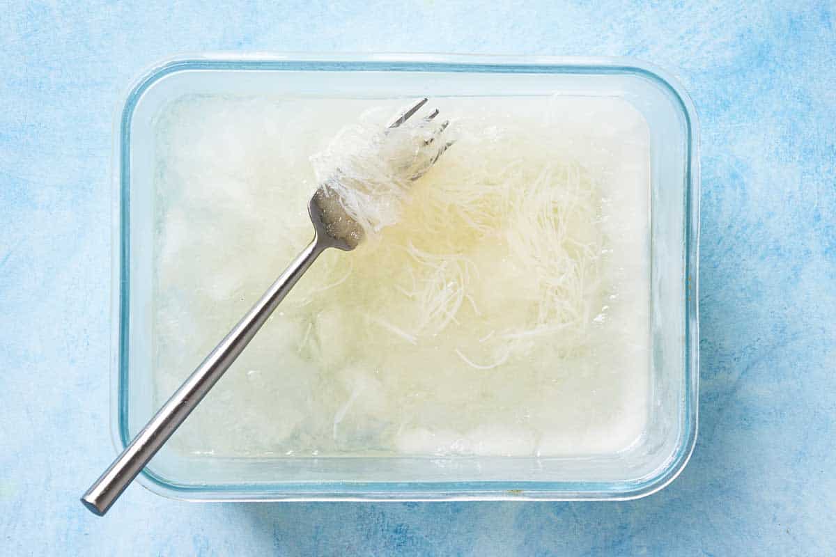 An overhead photo of the cooked and cooled vermicelli noodles mixed with the rose syrup in a freezer safe dish with a fork.