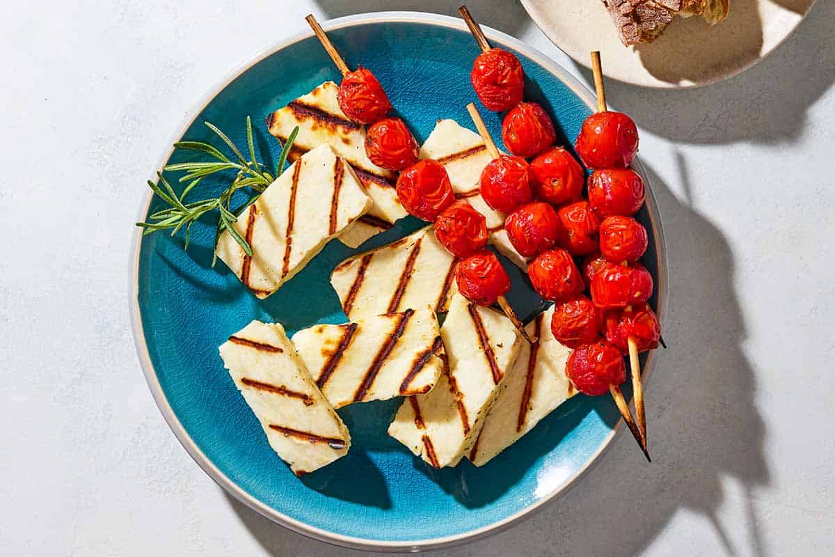 An overhead photo of grilled halloumi with blistered cherry tomatoes and sprigs of rosemary on a serving platter next to a plate with crusty bread.